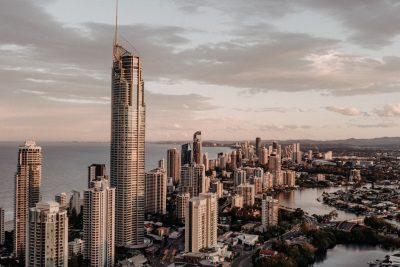 A drone photo of the Gold Coast skyline with skyscrapers, with one tall building in front of it and an ocean view behind it, golden hour lighting, cinematic, editorial photography, professional color grading, shot in the style of Hasselblad X2D. --ar 128:85