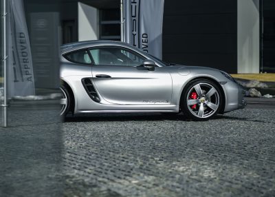 203 Porsche Cayman S in silver, parked on concrete near a Porsche retailer store with "ADVclientId Julien" written next to it. Shot in the style of canon eos r5, wide angle shot, high resolution photography. --ar 128:91