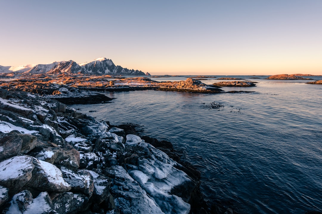 View of the ocean in Lofoten, Norway with snow-covered rocks and mountains on the horizon, golden hour lighting, Sony Alpha A7 III, 24mm f/8 lens, f/5.6 aperture, ISO speed 300, soft natural light, rule of thirds composition. –ar 128:85