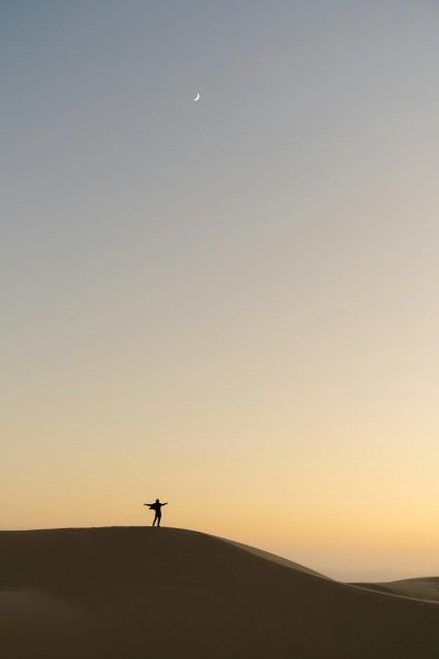 A lone dancer on the top of sand dune at dawn, with clear sky and crescent moon in background, symbolizing solitude and self vwew, minimalist photography style, soft lighting, wideangle lens, neutral tones, serene atmosphere, tranquil mood. --ar 85:128