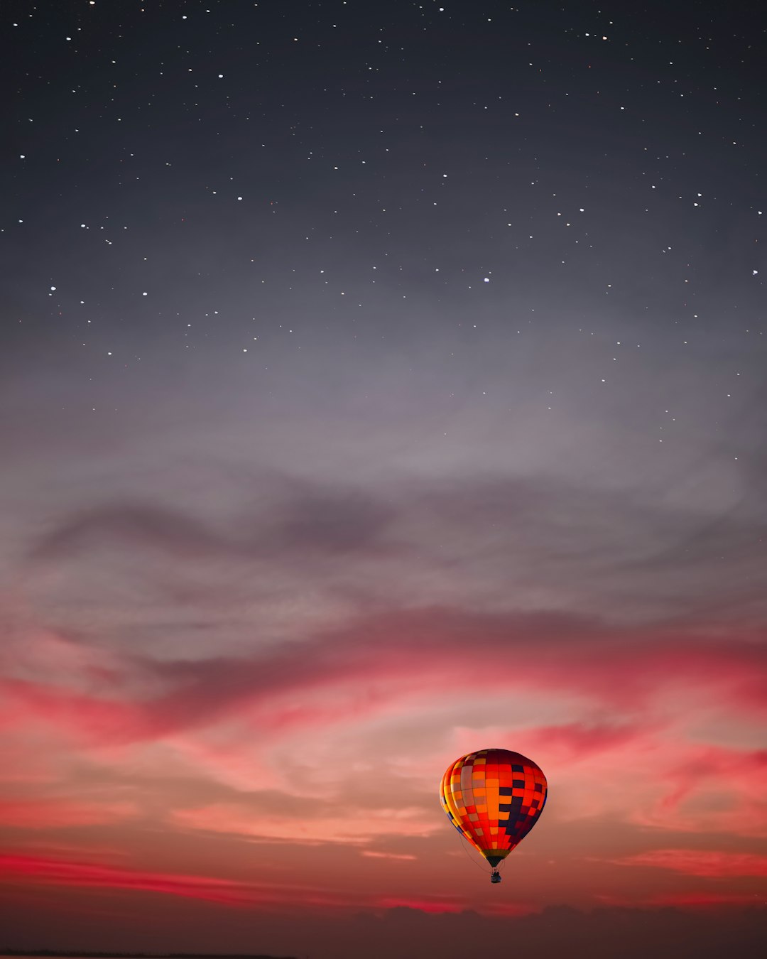 A hot air balloon floating in the sky at dusk, with stars and clouds creating an ethereal atmosphere. The balloon is colored red or pink against the twilight background, adding contrast to its silhouette. Shot in the style of Nikon D850 camera using f/4 lens. –ar 51:64