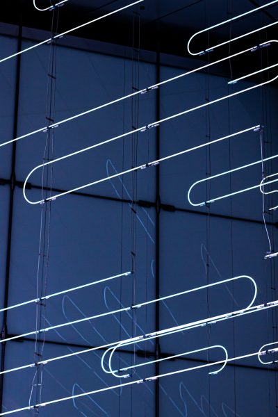 A series of white neon tubes are suspended in the air, creating an abstract pattern on dark blue glass walls. The lights form spirals and curved lines that resemble musical notes or arrows pointing upwards. This artwork creates a sense of movement and depth within the space in the style of musical notes or arrows pointing upwards. --ar 85:128