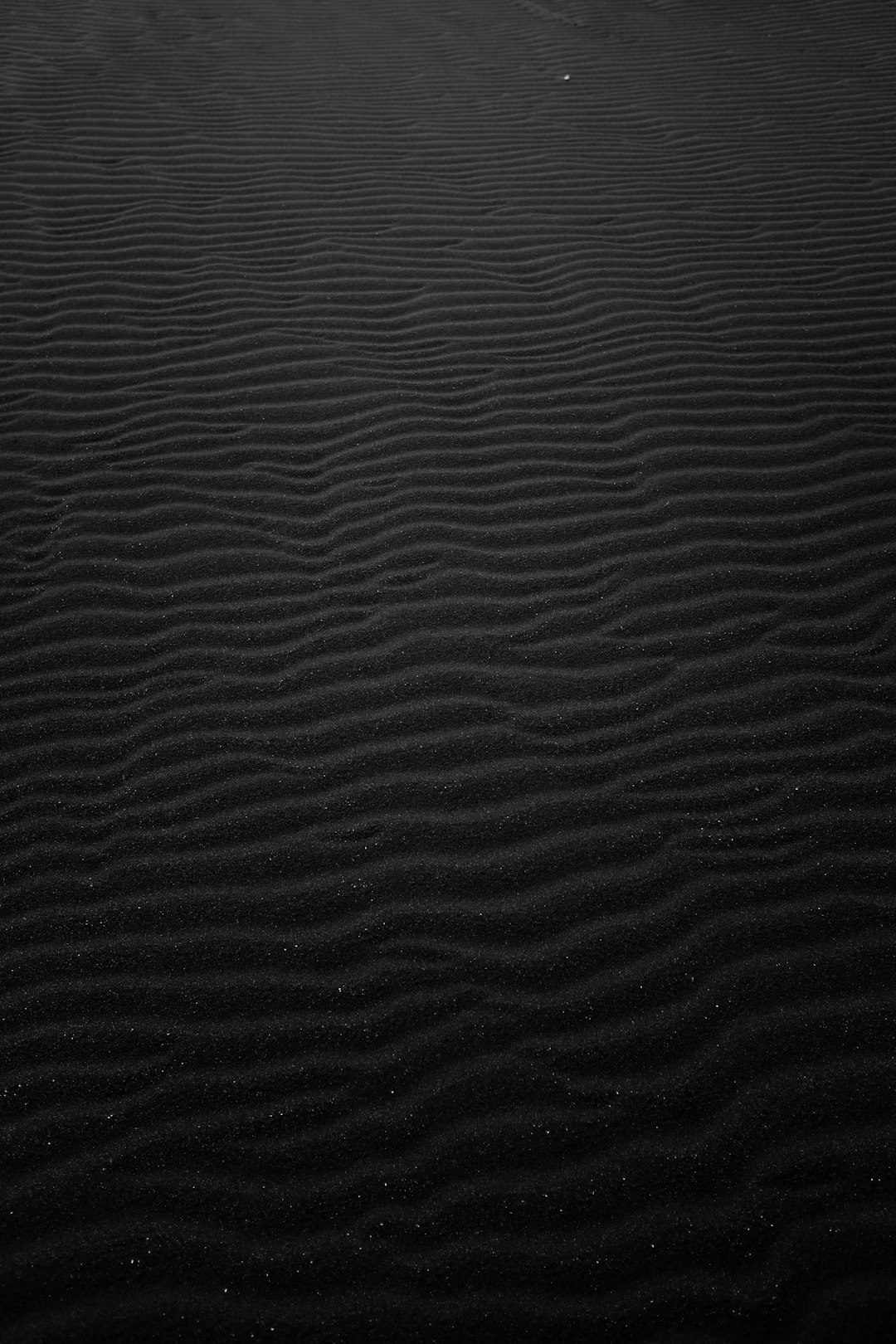 Black sand ripples in the desert, with a dark background and textured look, in black and white with high contrast, in a minimalist style, captured with a wideangle lens at night, as wind blows over sand dunes, with no people or objects around. The scene is in the style of minimalism. –ar 85:128