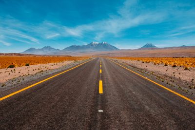 A long road leading to the Andes Mountains in Chile, a symbol of adventure and exploration. The desert landscape under blue sky creates an atmosphere for freedom or travel, symbolizing journeying into uncharted lands. In front there is two yellow lines on asphalt with mountains far away. --ar 128:85