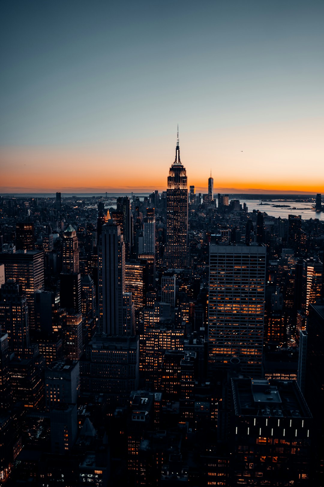 New York City skyline at dusk, capturing the city lights and skyscrapers from above. The view includes buildings like the Empire State Building and a drone shot to capture the full urban landscape. A dark blue sky with orange hues of sunset in the background. Shot on a Canon EOS R5 camera with an RF 24-70mm f/8 lens. –ar 85:128