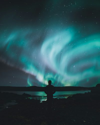 A person standing on the shore of an ocean, gazing at the vibrant aurora borealis in the starring night sky. The northern lights dance across the dark canvas with hues like green and blue, creating mesmerizing patterns that enhance their beauty. A man stands tall against his silhouette as he watches this celestial display. He is dressed casually yet elegantly to match the magical atmosphere. Captured from behind him using a wide-angle lens in the style of Nikon D850 DSLR camera with a hip level shot, high resolution, natural lighting, HDR. --ar 51:64