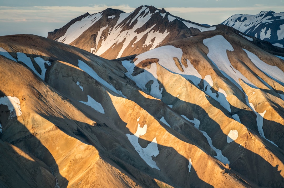 A high-resolution photo of the majestic and rugged landscape of Iceland’s Landmaar, showcasing golden sandstone mountains with snowcapped peaks, captured in the golden hour light. The photo showcases intricate patterns on the mountain side, with shadows cast by sunbeams creating an ethereal effect. Shot from above to capture the grandeur of these unique terrain features. –ar 32:21