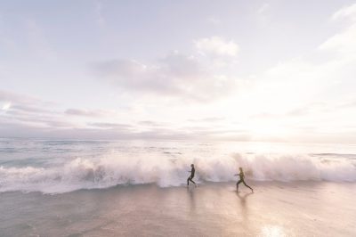 A wide shot of two people running on the beach, waves crashing in front of them, soft pastel colors, sunrise, minimalist photography, natural light, cinematic, captured in the style of Hassleblad X2D camera and lens --ar 128:85