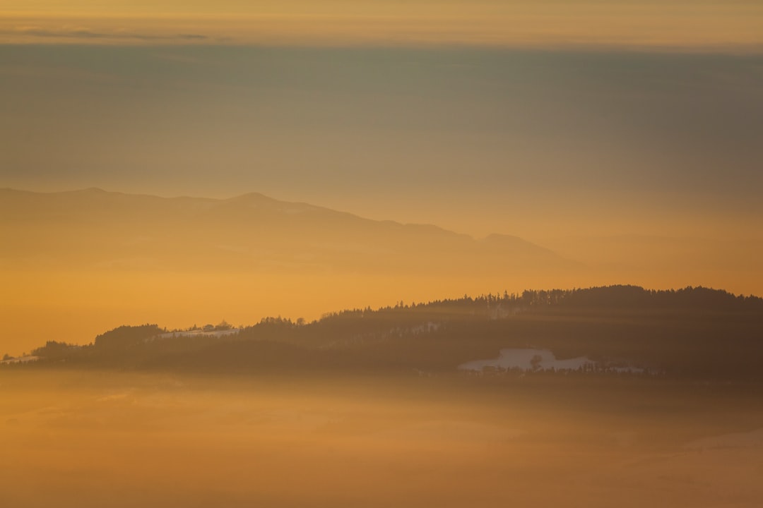 A golden sunrise over the mountains, with mist rolling in from below and distant forests visible through it. The sky is a gradient of orange to yellow, creating an ethereal atmosphere. In front of them lies a large plain covered by snow, with hills rising behind that can be seen faintly against the backdrop of dawn. This photo was taken using Canon EOS R5 camera. –ar 128:85