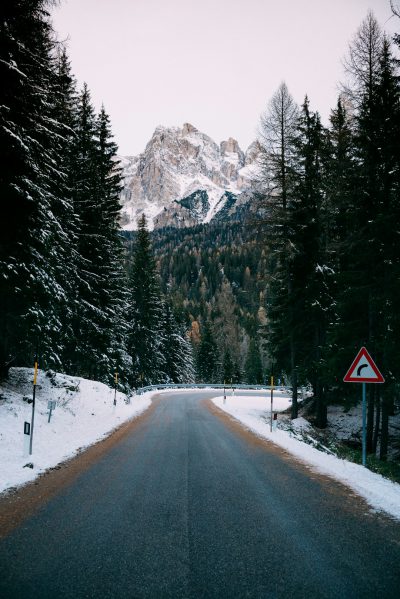 photo of an Italian road in the Dolomites, with snow on the ground and trees, a traffic sign visible on the right side, in the style of unsplash photography --ar 85:128