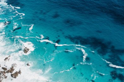 Aerial view of turquoise ocean waves and white foam, clear blue water, rocky shore in the distance, sunlight reflecting off surface, high resolution photography, stock photo, unsplash style --ar 128:85