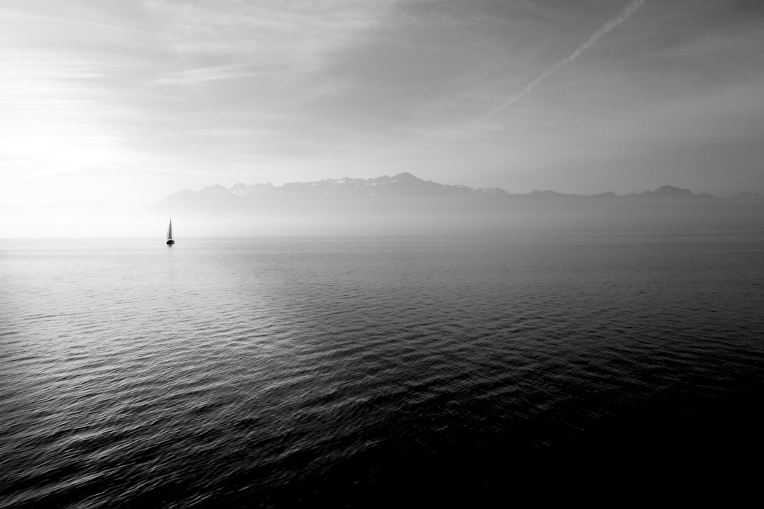 A black and white photograph of the swiss alps in the distance, with lake constance. In the foreground is an open sea with no land visible on its horizon. A small sailboat can be seen far away in the water. The sky above has low clouds and mist over it. There’s almost nothing but water in sight. The overall mood is calm and serene. It feels like you’re looking at something that looks peaceful yet lonely. –ar 128:85