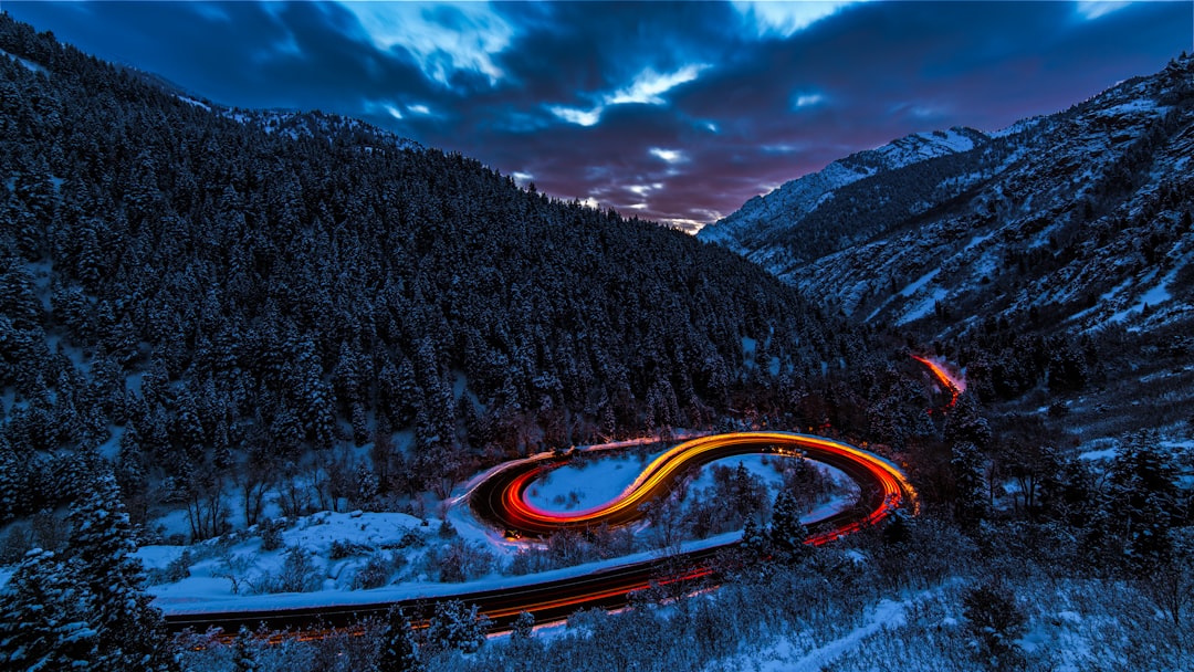 A long exposure photograph of cars’ tail lights creating an artistic swirl on the winding road in winter mountains with snow-covered forest at twilight, creating a captivating and mysterious scene. The sky is dark blue with clouds, adding depth to the composition. This photo was taken using a Sony A7R IV camera, capturing the dynamic light trails and the serene beauty of nature’s landscape in the style of minimal editing. –ar 16:9