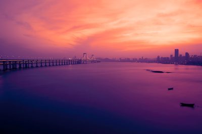 Wencun Bridge, the vast sea under the twilight sky in springtime, distant view of the Shenzhen city skyline, distant boats on the water surface, purple and orange color tone, wideangle lens, long exposure photography style, tranquil and mysterious atmosphere. --ar 128:85