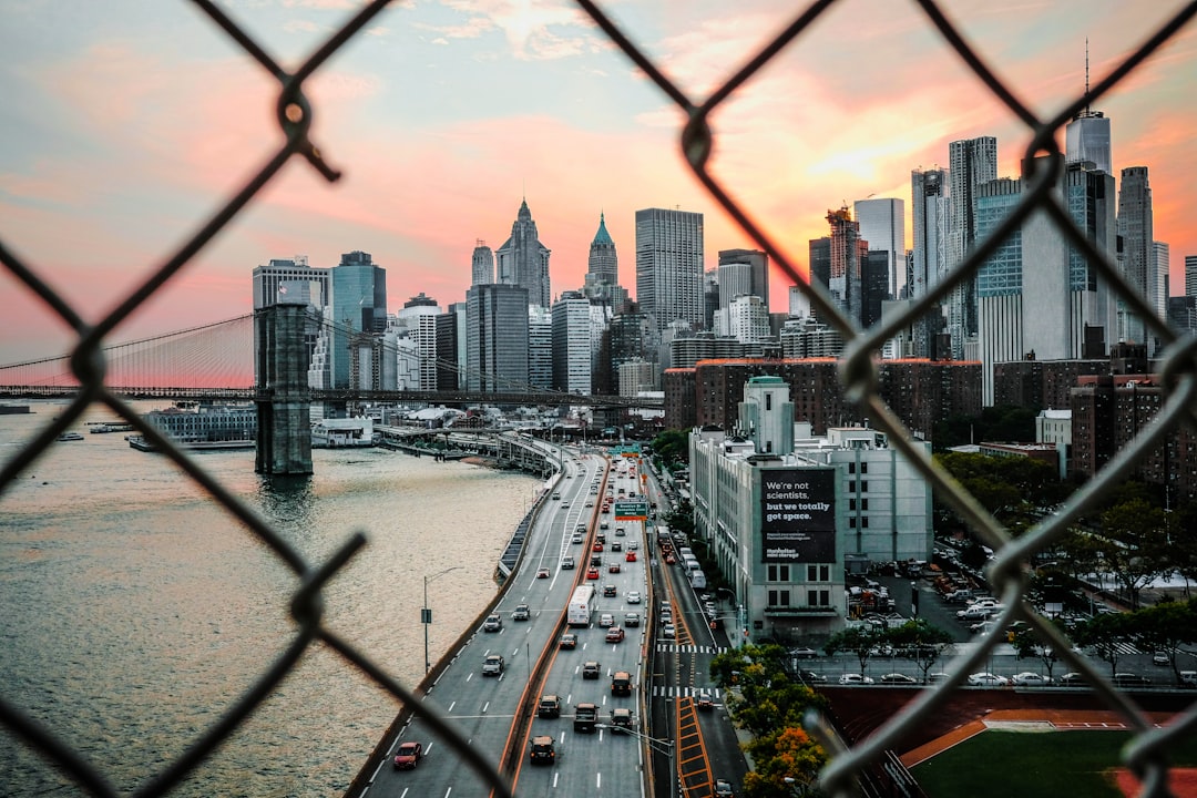 View of the city skyline from behind an old chain link fence, cars on bridge in the foreground, New York City skyline, New York cityscape, sunset, New York City buildings and streets, buildings in the background, photography, city view through the wire mesh. –ar 128:85