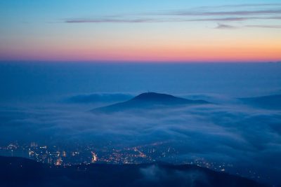 view from the top of vAfter pol现d, the city lights below and fog rolling over mountain at dawn, Chodsko national park, captured with Sony Alpha A7 III, f/8 aperture setting, and ISO 200 for balance in blue hues and warm tones. The sky is painted in the style of soft pastel colors of pink, orange, purple, and indigo. There's a thin layer of clouds between meadow hills. In front there can be seen silhouette of Keshet Mountain. On its peak stands television tower of wrapped around it mist. --ar 128:85