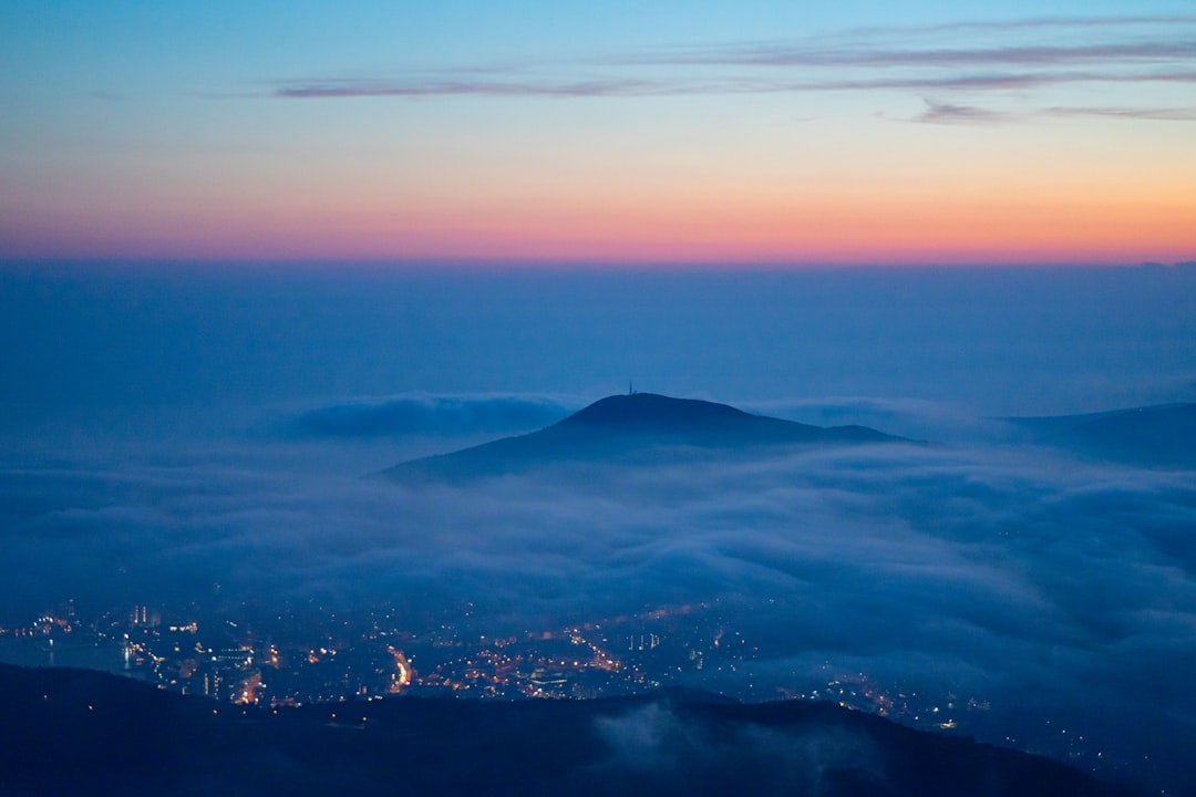 view from the top of vAfter pol现d, the city lights below and fog rolling over mountain at dawn, Chodsko national park, captured with Sony Alpha A7 III, f/8 aperture setting, and ISO 200 for balance in blue hues and warm tones. The sky is painted in the style of soft pastel colors of pink, orange, purple, and indigo. There’s a thin layer of clouds between meadow hills. In front there can be seen silhouette of Keshet Mountain. On its peak stands television tower of wrapped around it mist. –ar 128:85