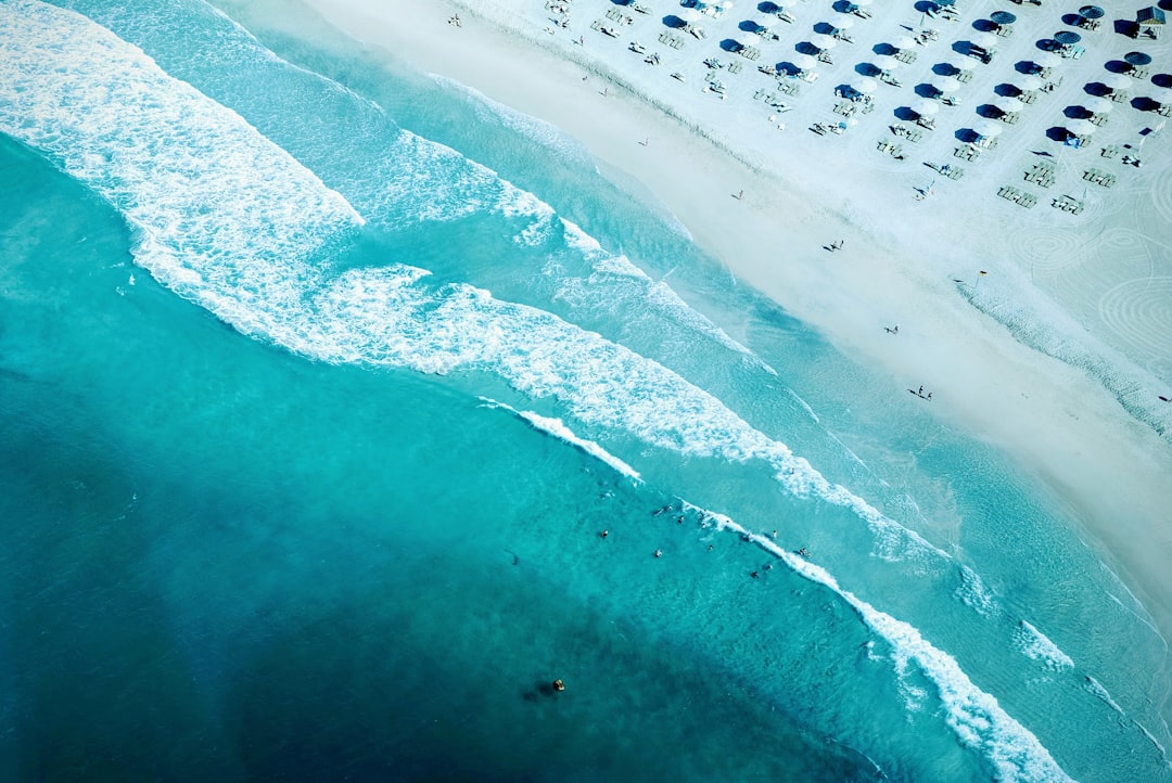 Aerial view of the turquoise ocean waves and white sandy beach with umbrellas in Florida, United States. The water is a clear blue with people swimming or surfing on surfboards. A sunny day at the Miami Beach. View from above. High resolution photography, high definition, ultra detailed. –ar 128:85