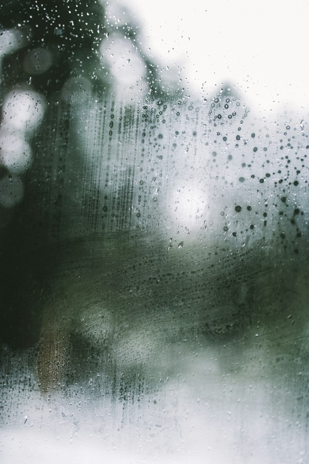 photograph showing raindrops on a window with a blurry background, water drops, foggy background, soft focus, depth of field blur, double exposure effect, overcast cloudy sky visible, green trees in the distance, grainy film filter effect, white background, muted colors, texture details, wet surface, raindrops –ar 85:128