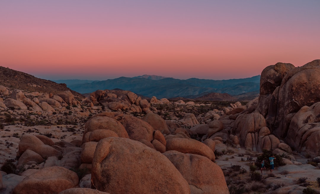 Boulders at Joshua Tree National Park during sunset, with mountains in the background, people hiking, and a pink and purple sky. High resolution landscape photography of the scene taken with a Canon EOS Mark III 50mm f/2.8L II USM lens, including a panoramic shot. –ar 128:77