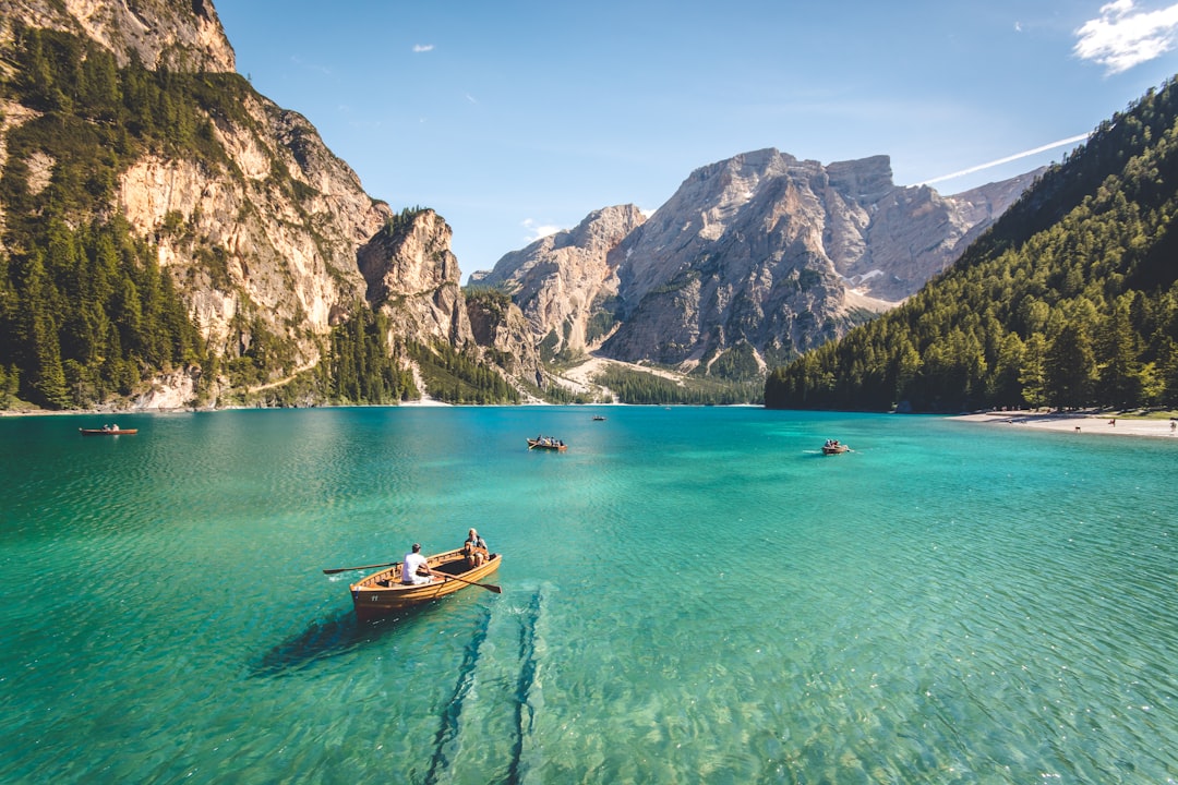 A stunning view of the Pragserwaz Lake in Dolomites, Italy with clear turquoise waters and people rowing boats on it. The background features majestic mountains covered by dense forests and some beach areas, adding to its beauty. In one part there is also visible an old wooden boat floating on water. This picturesque scene captures nature’s grandeur and elegance, making for a captivating travel photograph. –ar 128:85