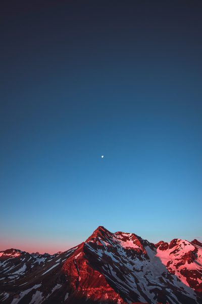 a mountain with snow and red glow, clear blue sky, moon in the distance, photograph by unsplash --ar 85:128