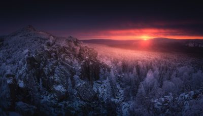 Sunrise over the snowy mountains of Z.brno, Czech Republic, with snow-covered rocks and forest in the foreground. A beautiful winter landscape captured in the style of a professional photographer at dawn. Wide angle view from top to bottom. The sky is painted red as if it were a canvas, while the sun rises on the horizon. This scene creates an atmosphere of tranquility and beauty, emphasizing nature's grandeur. Shot using a Nikon D850 camera, wide-angle lens, f/4 aperture, ISO 230, shutter speed 6 seconds. --ar 128:73