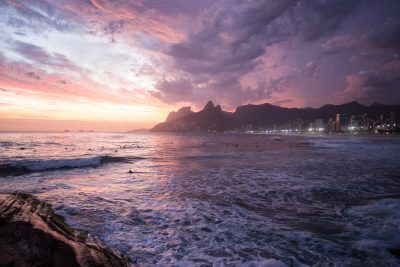 A photo of Ipanema beach at sunset, with a pink and purple sky, surfers in the water, mountains in the background, buildings and lights, beautiful clouds, dramatic and cinematic, with a 35mm lens at f/20, 86k. --ar 128:85