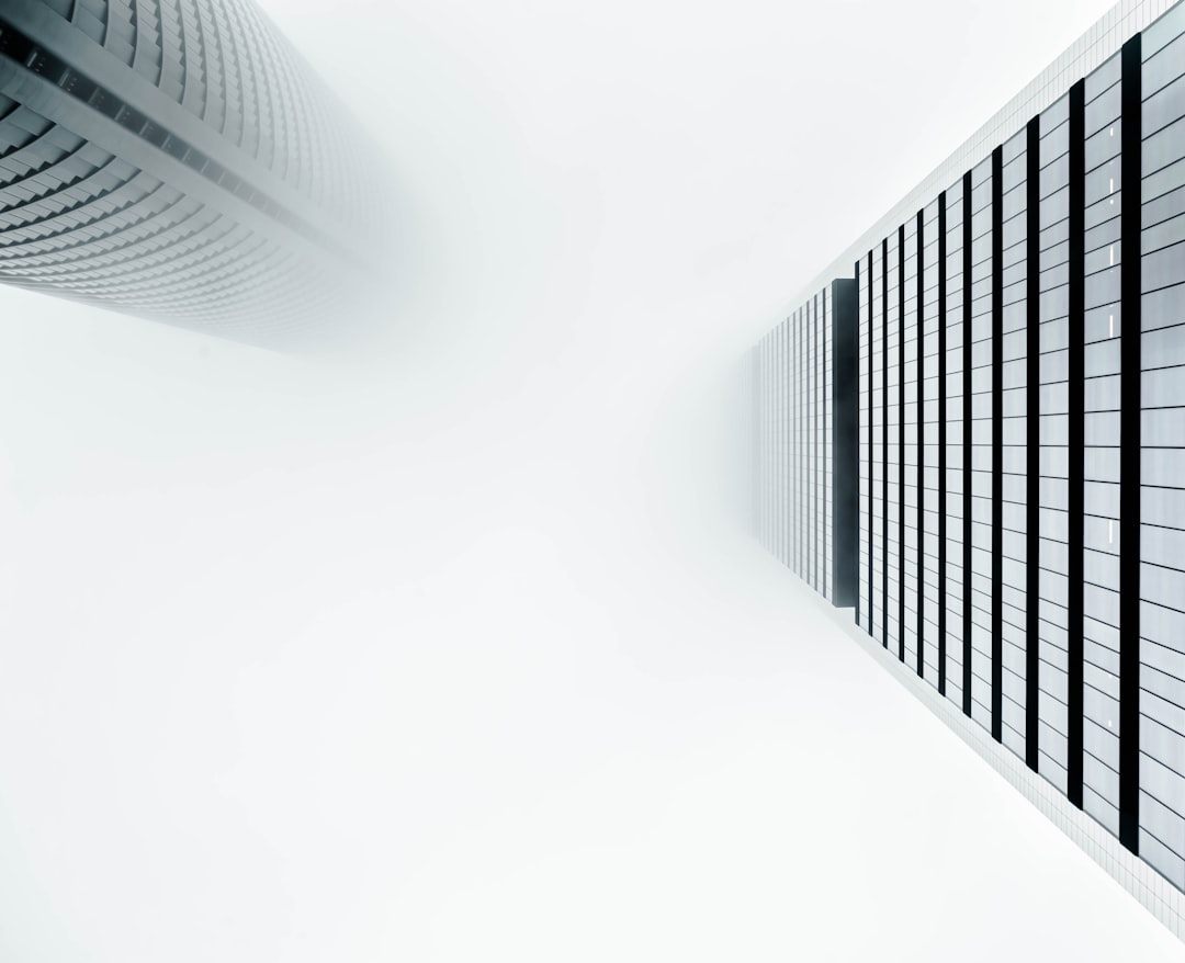 A white background with two modern skyscrapers on the sides, one slightly behind and partially obscured in fog, captured from an upward perspective. The sky is clear above them, creating a sense of depth in the scene. Soft lighting highlights their sleek lines against the bright backdrop. This composition creates contrast between lightness and heavy structure, symbolizing city life. –ar 16:13