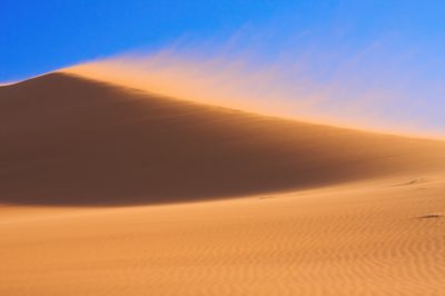 A sand dune in the desert with a blue sky background, captured in the style of Canon EOS R5 using an f/8 aperture for detail and depth. The wind blows up dust clouds around it, creating dynamic patterns of light and shadow on its surface. --ar 128:85
