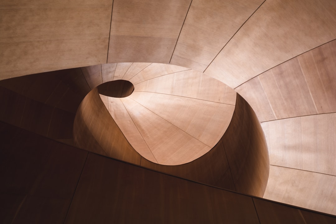 An architectural photograph of an undulating interior wall made from timber, designed in the style of [Zaha Hadid](https://goo.gl/search?artist%20Zaha%20Hadid). The lighting is soft and natural, highlighting the fluid lines and organic shapes that characterize her architecture. In one corner there’s a circular hole through which you can see part of another room with light coming through it. This photo was taken using a Canon EOS R5 camera with a Canon RF80mm f/2 lens. –ar 128:85