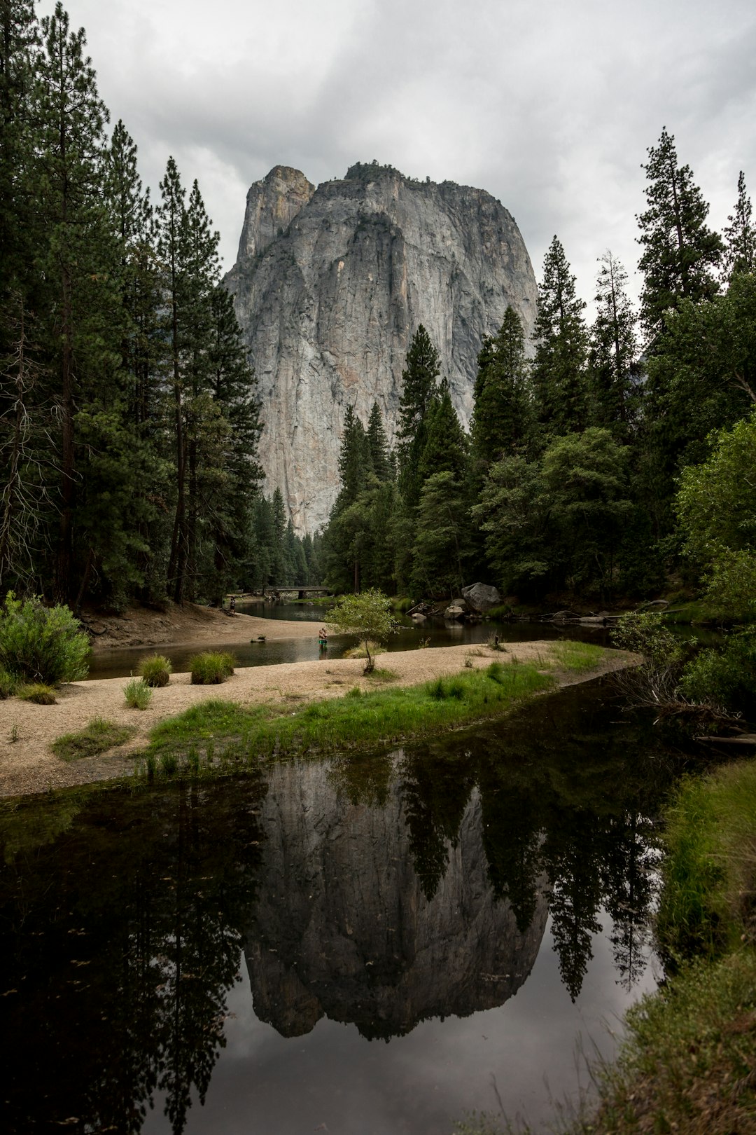 El Capitan in Yosemite National Park, reflecting on the water of the pond at its base, surrounded by dense forest and flat ground covered with sand. The sky is overcast, adding to the natural beauty of the scene. –ar 85:128