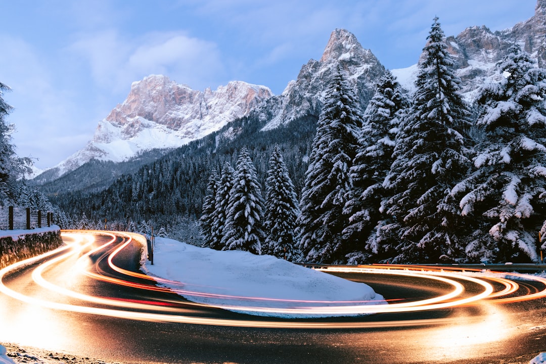 long exposure photo of cars driving around the hairpin turns on the Dolomites in winter, snow covered mountains and trees in the background. The photo is in the style of blurred lights from the cars turning the curves on the mountain road while the snowy landscape remains still. –ar 128:85