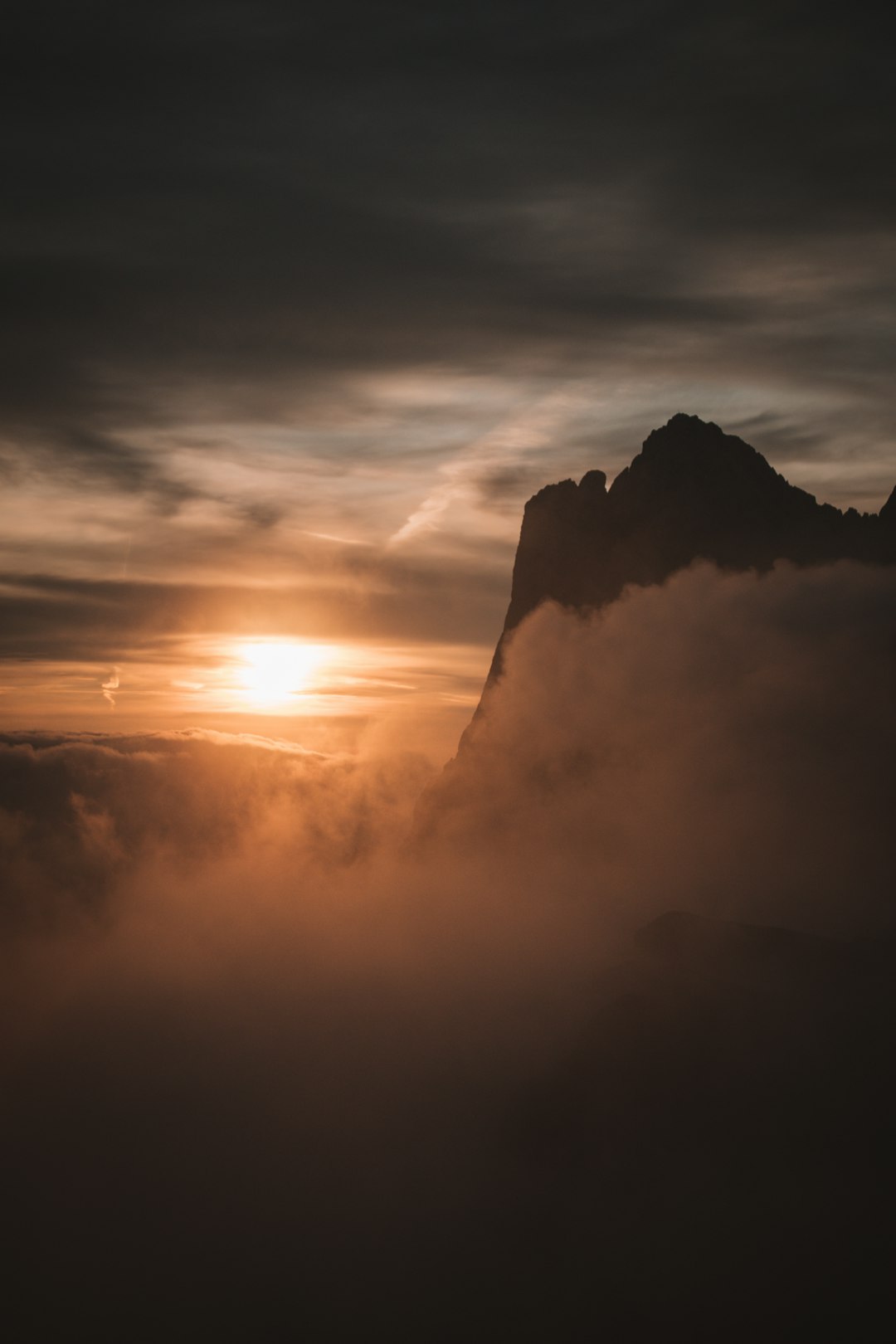 The sun rises behind the mountain, the clouds cover some of its silhouette which is visible, there is fog below it, in nature, dark tones are used, high resolution, captured in the style of Canon R5. –ar 85:128