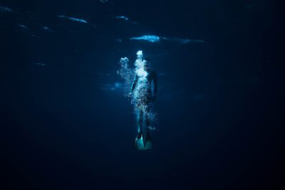 Full body shot of an ethereal woman wearing flippers, swimming in the deep dark ocean. The lighting is dim and cinematic. Shot with a Sony Alpha A7 III camera using a wide angle lens to capture the vastness around her. --ar 128:85