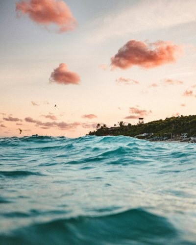 photograph of the sea in Tulum, Mexico with pink clouds and kitesurfers on it, shot from inside an ocean wave looking towards a tropical island with buildings, shot at golden hour, beautiful sky, taken in the style of Sony Alpha A7 III camera with f/8 aperture setting, 35mm focal length lenses, natural light, soft focus, bokeh effect, minimalistic, neutral color tones, no contrasty colors, calming effect, --ar 51:64