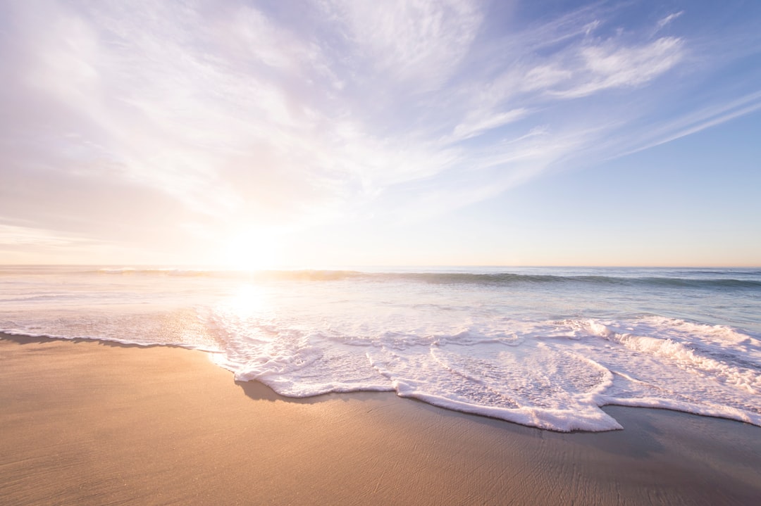 Photograph of a beach with beautiful waves and sand, during sunrise, with a sky full of white clouds, over a calm sea, on a sunny day, with a light blue background, during the golden hour, with a peaceful, calm and relaxing atmosphere, in the style of nature photography, taken with a canon eos r5 camera. –ar 128:85