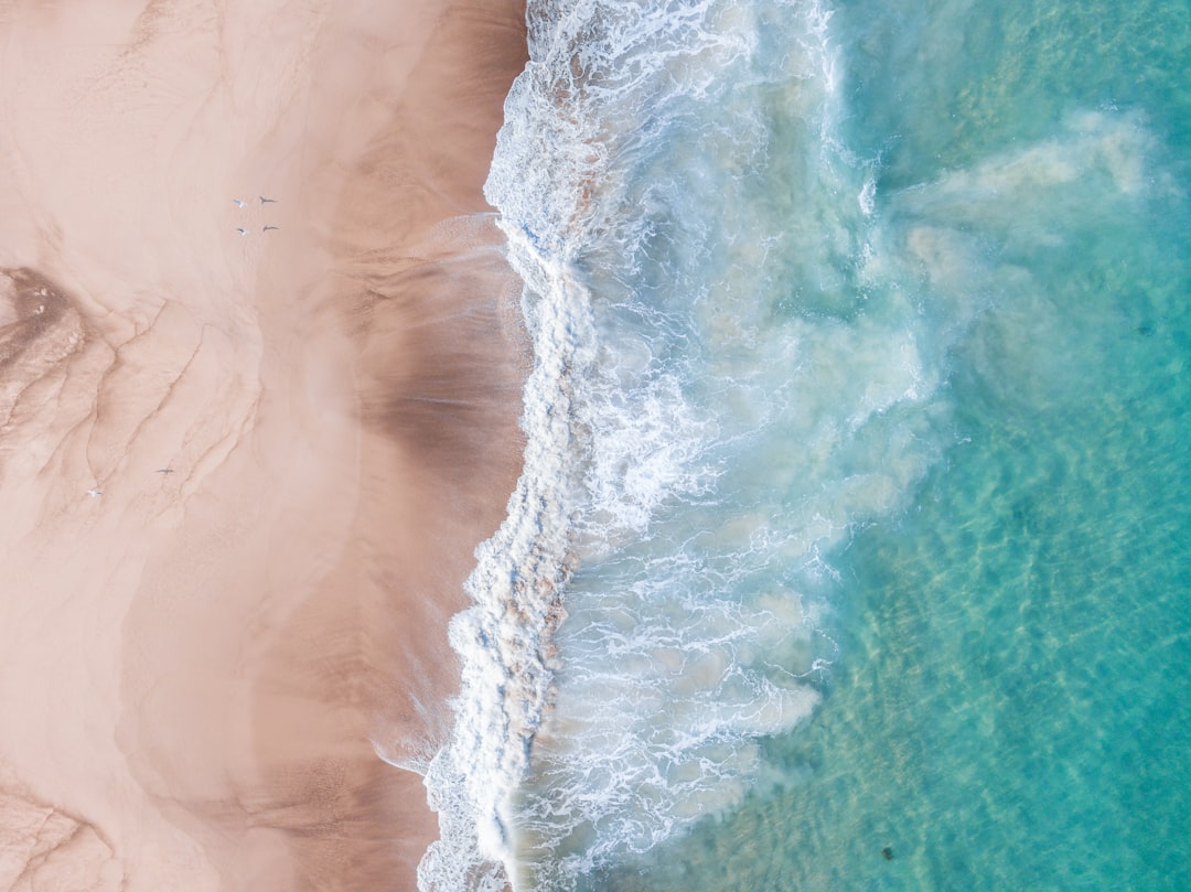 Beautiful aerial view of the sandy beach and turquoise sea water, with waves crashing onto shore. The shot is taken from above in the style of an drone, creating a stunning and breathtaking scene. This photograph was captured using a high-resolution camera to showcase every detail in exquisite clarity and color depth. It has been carefully arranged for easy presentation on social media or other digital platforms. –ar 128:95