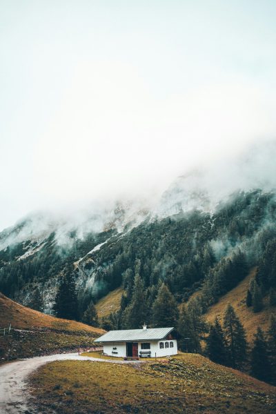 A small white house on the side of an alpine mountain, surrounded by dense forest and misty clouds, captured in a minimalist photographic style with a focus on natural beauty and serenity. The scene is captured from a low angle to emphasize the cabin's architecture against nature's backdrop, in the style of minimalist photography. --ar 85:128