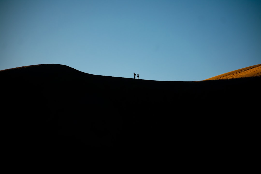 A silhouette of two people walking on the top of sand dunes, with a clear blue sky in the background. The contrast between dark and light creates depth to the scene. A sense of adventure is captured as they explore the vastness under the open air. Photographed in the style of David Bur signature, professional photographer. –ar 128:85