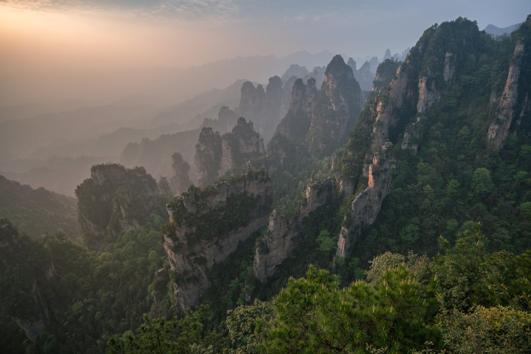 Zhangjiajie National Forest, hazy mist envelops the towering peaks and lush greenery, creating an otherworldly landscape that is both awe-inspiring and serene. The rugged rock formations of Zhangji Fairy Mountain stand tall against the backdrop of morning light, casting long shadows over the dense forest below. Shot with a Sony A7R IV camera using a G Master FE lens at f/8 aperture for sharp focus on the rocks in the style of Sony. –ar 128:85