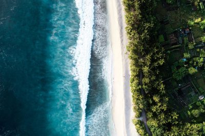 An aerial view of a white sandy beach and blue ocean, with waves breaking on the shore and palm trees. In the style of unsplash photography. --ar 128:85