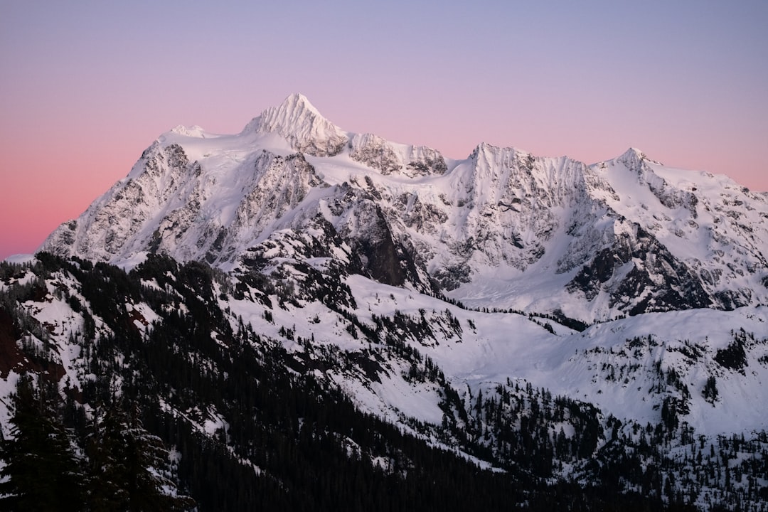 A snow-covered mountain peak at sunset, Shuksan Mountain captured in the style of medium format film grain, with a pink and purple sky. Snowcapped peaks and a dense forest below the summit, shot with a Sony Alpha A7 III using a wide-angle lens at f/4 aperture to capture detail in the sunset lighting at high resolution. –ar 128:85