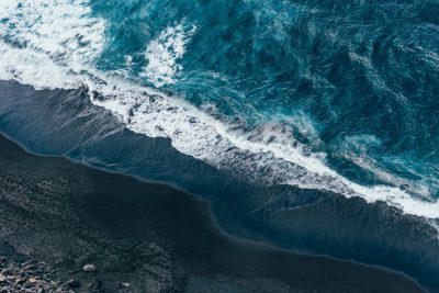 View from above of dark blue sea waves hitting the black sand beach, drone shot, photo realistic, hyperrealistic photography, in the style of cinematic grading, in the style of color grading. --ar 128:85