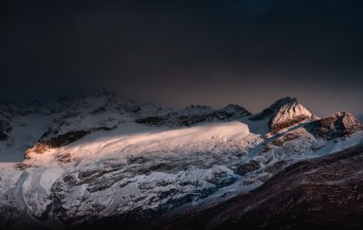 A snow-covered mountain range bathed in the soft glow of dusk, with dramatic lighting creating deep shadows and highlights that accentuate its rugged beauty. The mountains appear to rise high into an overcast sky, adding depth to the scene. This photo was taken using a Sony A7R IV camera in the style of dramatic lighting. --ar 128:81
