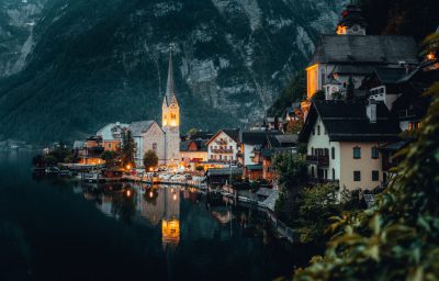 A photo of HallLEMmer in Austria, hall Citation view, evening time, buildings illuminated with lights, and the lake reflecting their beauty. The town is nestled at the foot of mountains, with charming architecture, and church steeple standing tall above it. This scene captures its picturesque charm and serene atmosphere. --ar 128:81