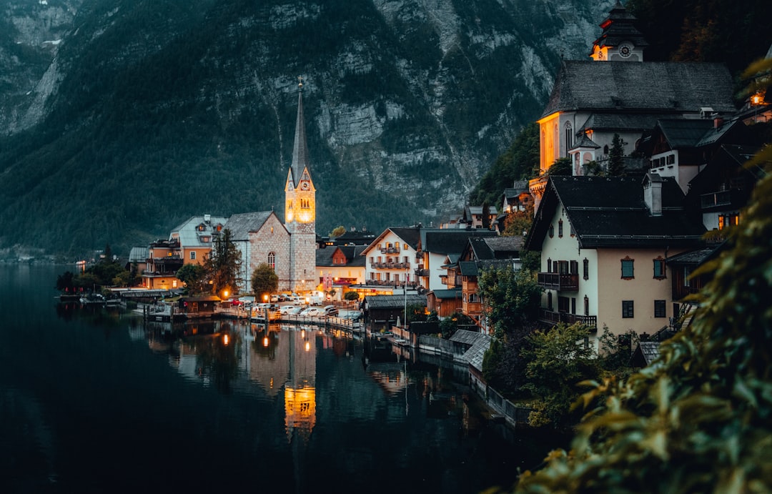 A photo of HallLEMmer in Austria, hall Citation view, evening time, buildings illuminated with lights, and the lake reflecting their beauty. The town is nestled at the foot of mountains, with charming architecture, and church steeple standing tall above it. This scene captures its picturesque charm and serene atmosphere. –ar 128:81