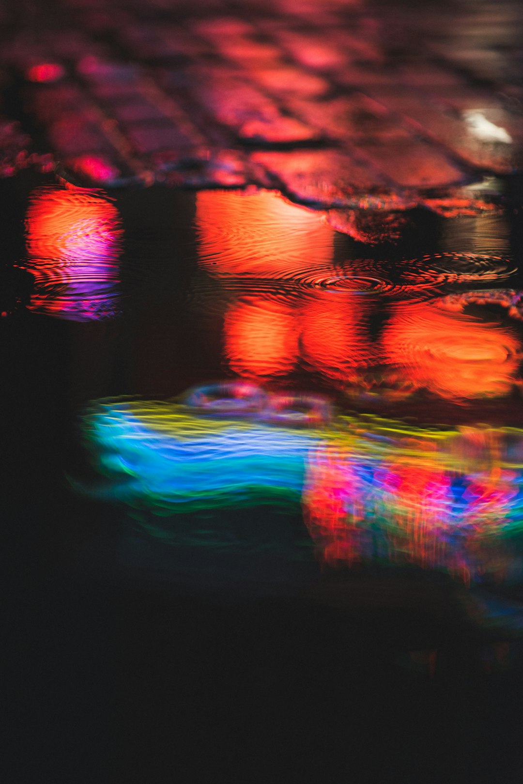 A blurry, colorful reflection of neon lights on a water surface, capturing the play between light and shadow. The reflections create an abstract composition that evokes warmth and joy in urban life. Shot with a Canon EOS R5 mirrorless lens at an f/2.8 aperture for a shallow depth of field. –ar 85:128