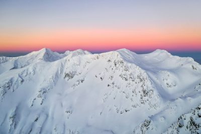 Aerial view of snow-covered mountains at sunrise, pink and purple sky, Sony A7R IV with panoramic lens, high resolution for crisp details, in the style of aerial photography, snowcapped peaks. --ar 128:85