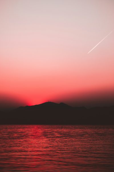 Red sky, distant islands and mountains, sunset, plane trail, calm sea surface, simple composition, minimalist style, wideangle lens, distant view, silhouette effect, soft light, tranquil atmosphere. in the style of minimalist. --ar 85:128